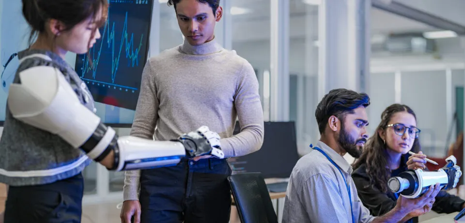 A robotics team develops a new bionic limb, inspecting the prototype while analyzing real-time performance data. A concept for digital twins, agile R&D, and mechatronics innovation.