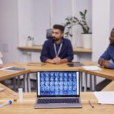 Brain scan on laptop screen on background of three young intercultural medical experts brainstorming and discussing new strategies