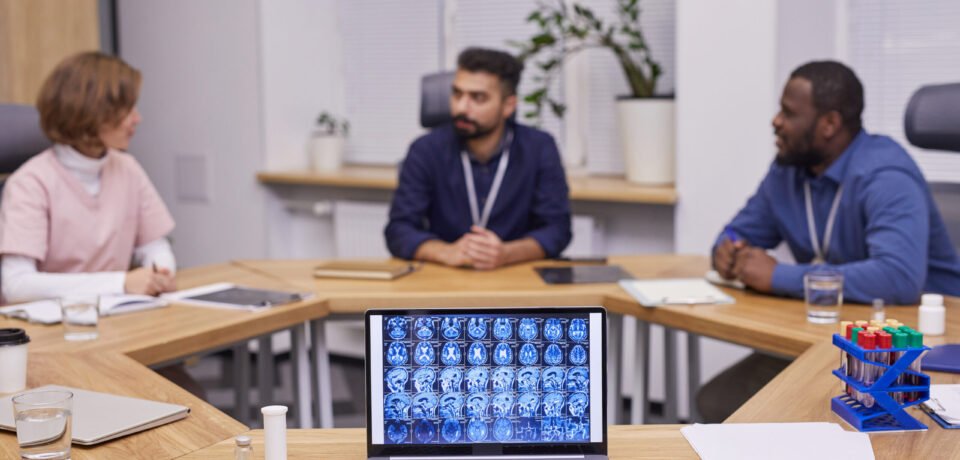 Brain scan on laptop screen on background of three young intercultural medical experts brainstorming and discussing new strategies