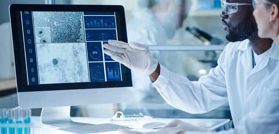 Colleagues working together in laboratory African male scientist pointing at computer monitor with micro image of bacterium and discussing it with his colleague