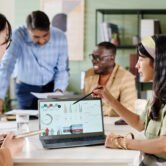 Multi-ethnic team discussing business analytics on laptops sitting around conference table, sharing ideas and analyzing data. Professional setting with open atmosphere and modern office design