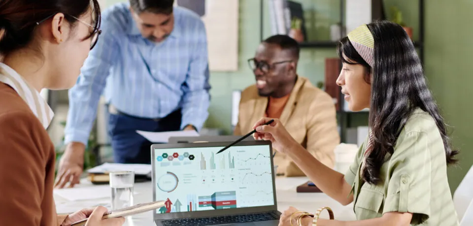 Multi-ethnic team discussing business analytics on laptops sitting around conference table, sharing ideas and analyzing data. Professional setting with open atmosphere and modern office design