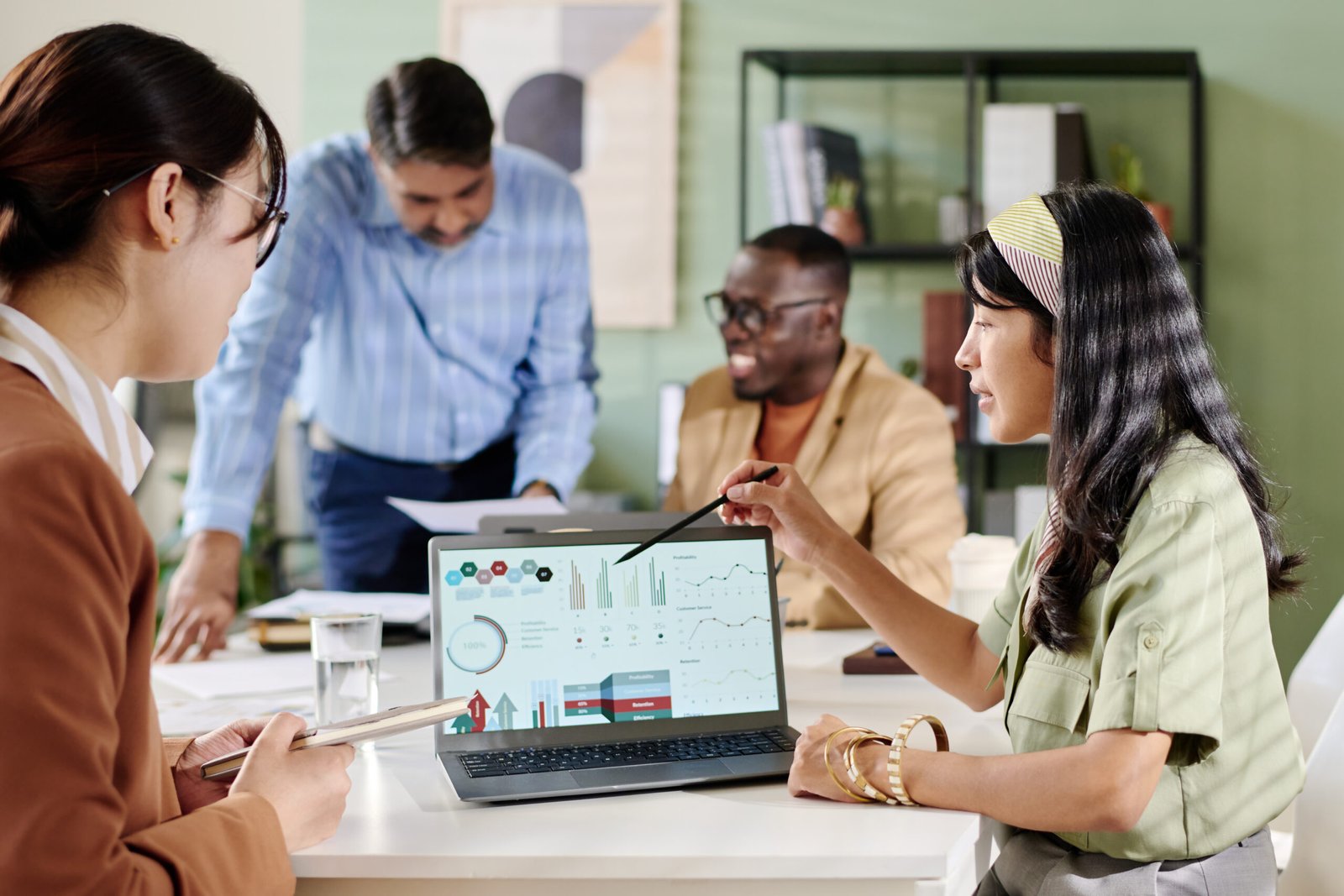 Multi-ethnic team discussing business analytics on laptops sitting around conference table, sharing ideas and analyzing data. Professional setting with open atmosphere and modern office design