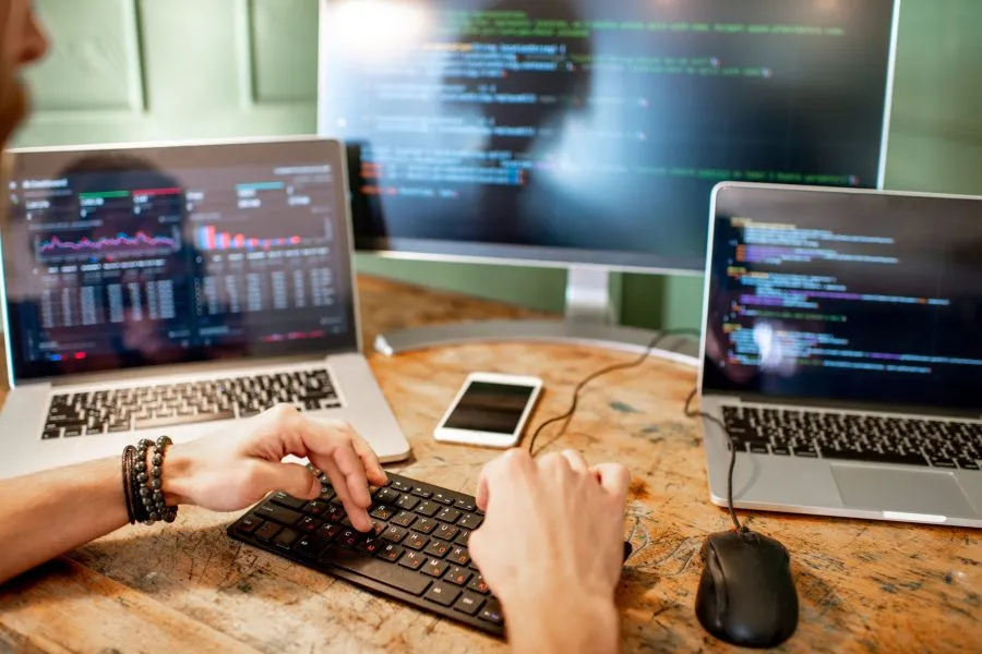 Young programmer writing a program code sitting at the workplace with three monitors in the office