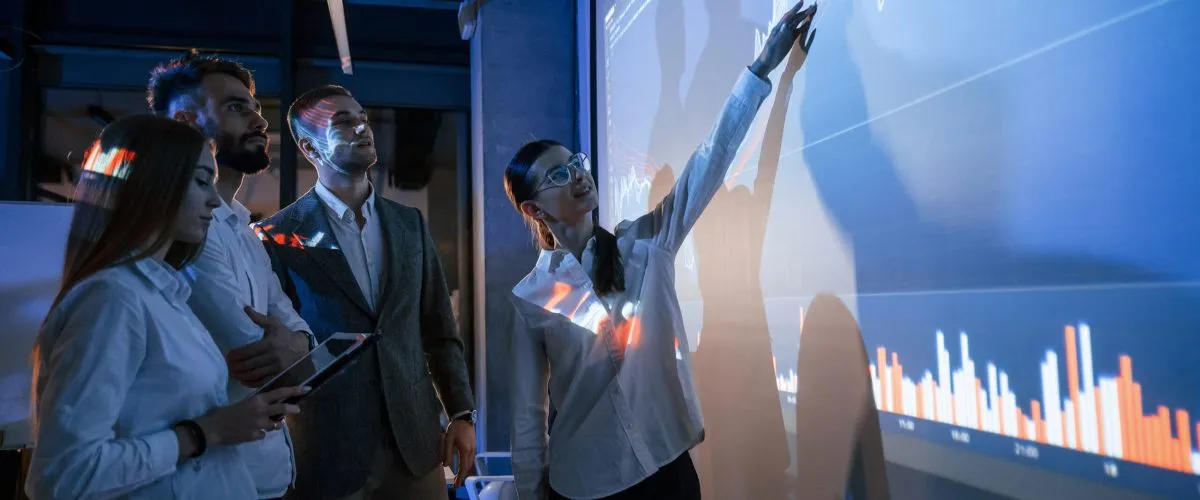 Holding tablet. Male leader talking to employees, showing the plan on the projector in office of stock exchange company.