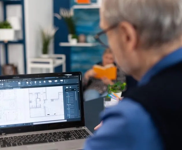 Overshoulder shot of senior architect working on laptop to finish a project. Experienced construction engineer designer looking at house blueprints on laptop computer from home office.