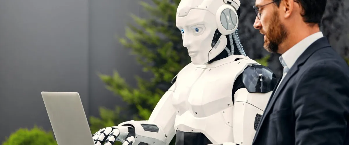 A male businessman in a business suit standing with a robot against the background of a building and using a laptop discussing work. The future with artificial intelligence.