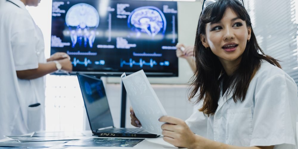 Team of female doctor check on scan results paper and looking at monitors in laboratory, share medical knowledge and experience benefits her coworkers and patients before surgery and treatment