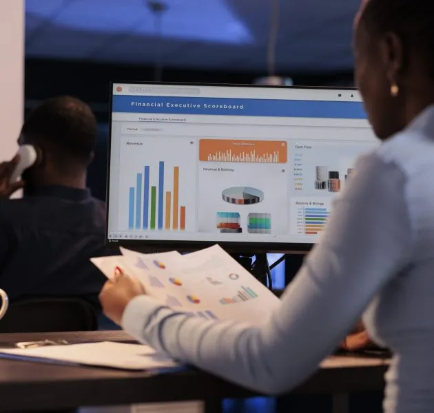 Young businesswoman working with statistics charts on computer monitor, analyzing company data on paperwork. Using online information to create research report before deadline, at night after hours.